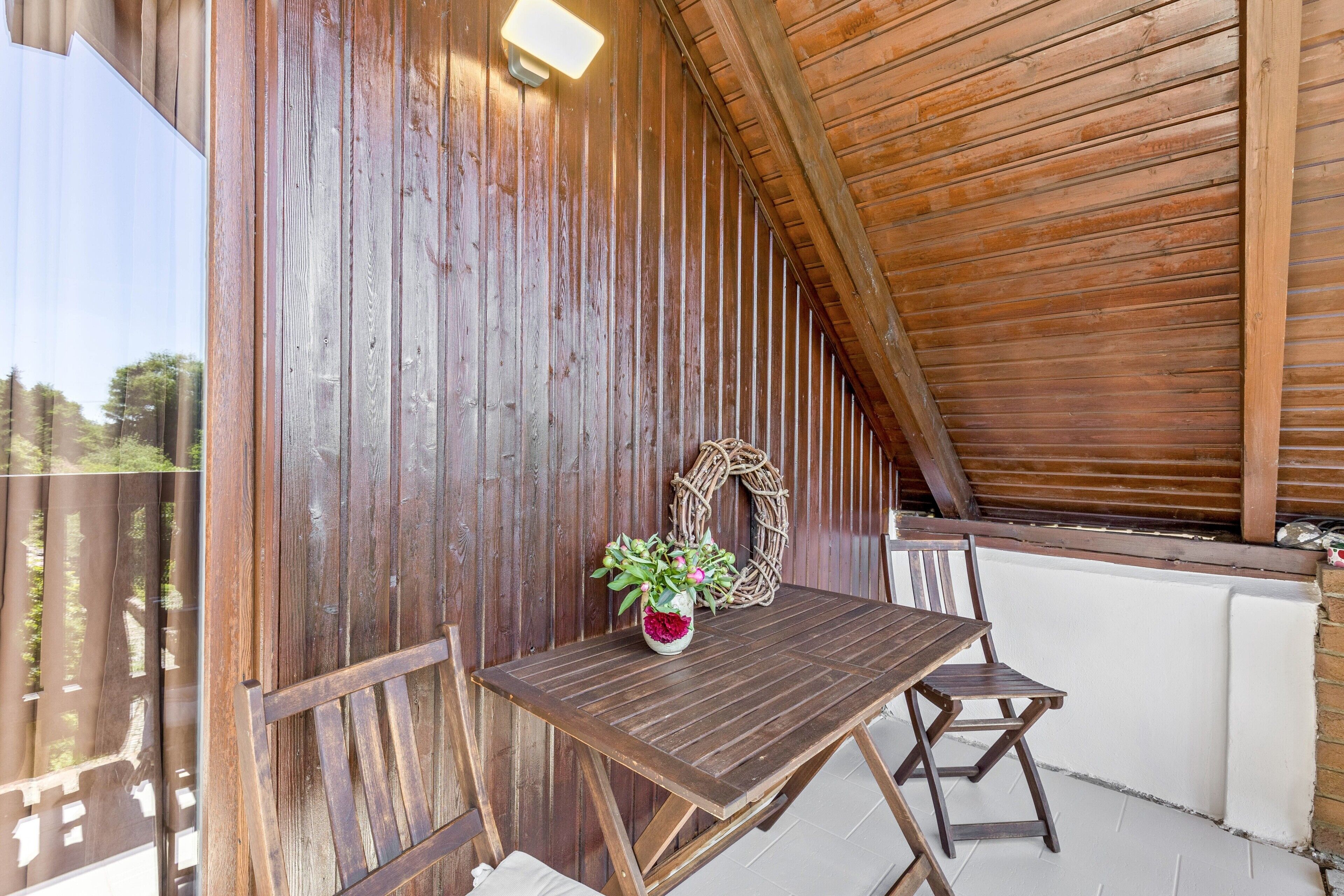 A wooden table with a woven wreath and a vase of flowers, a wooden bench, and a view of the outdoors.