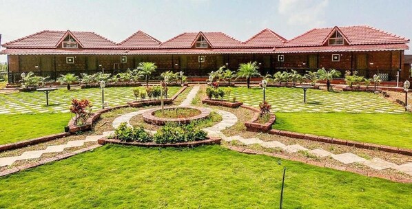 A building with a red-tiled roof and a courtyard with a central circular garden, surrounded by a low brick wall and palm trees. - Kabila Agro Tourism (Mahabaleshwar)