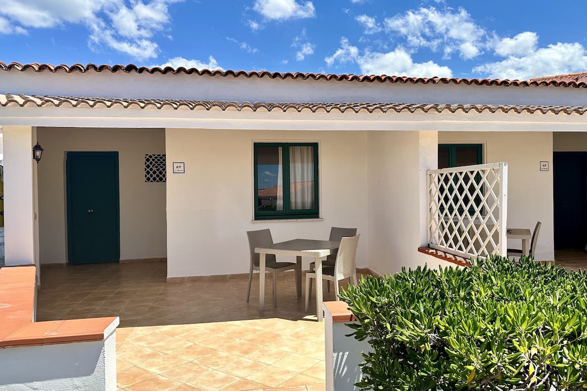 A two-story building with a tiled roof, a covered patio area with a table and chairs, and a small garden.