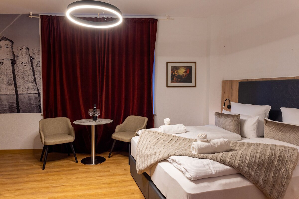 A hotel room with a bed, two chairs, a small round table, a painting, and a curtain.