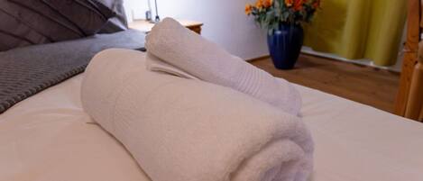 A neatly made bed with a rolled white towel, a dark grey pillow, and a vase with orange flowers on a wooden floor.