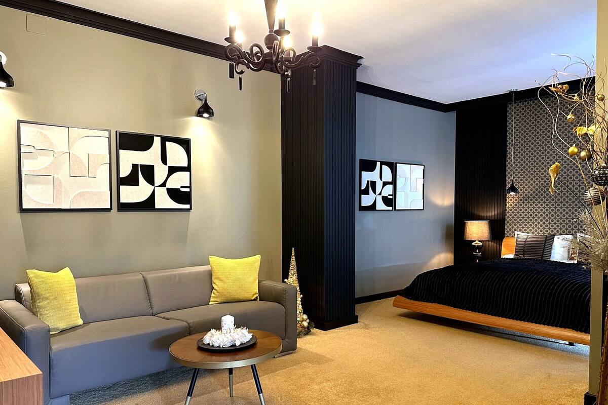 A modern hotel room with a grey sofa, a round coffee table, a bed with a dark headboard, and abstract wall art.