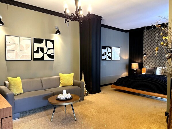 A modern hotel room with a grey sofa, a round coffee table, a bed with a dark headboard, and abstract wall art.