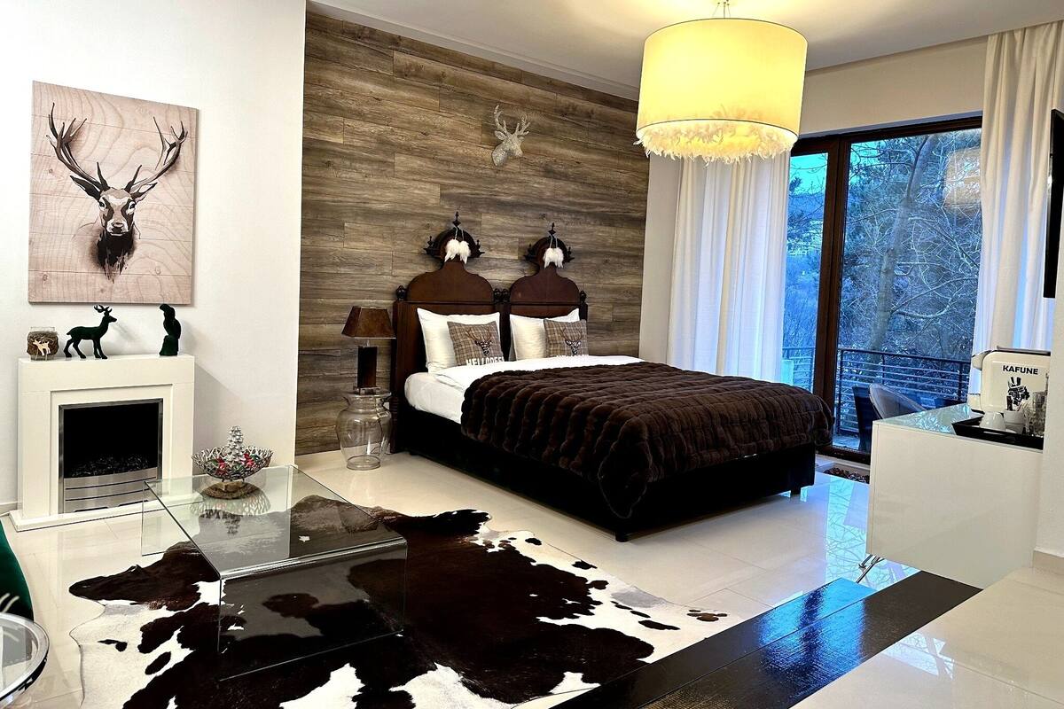 A modern bedroom with a bed, a chandelier, a fireplace, and a large window.