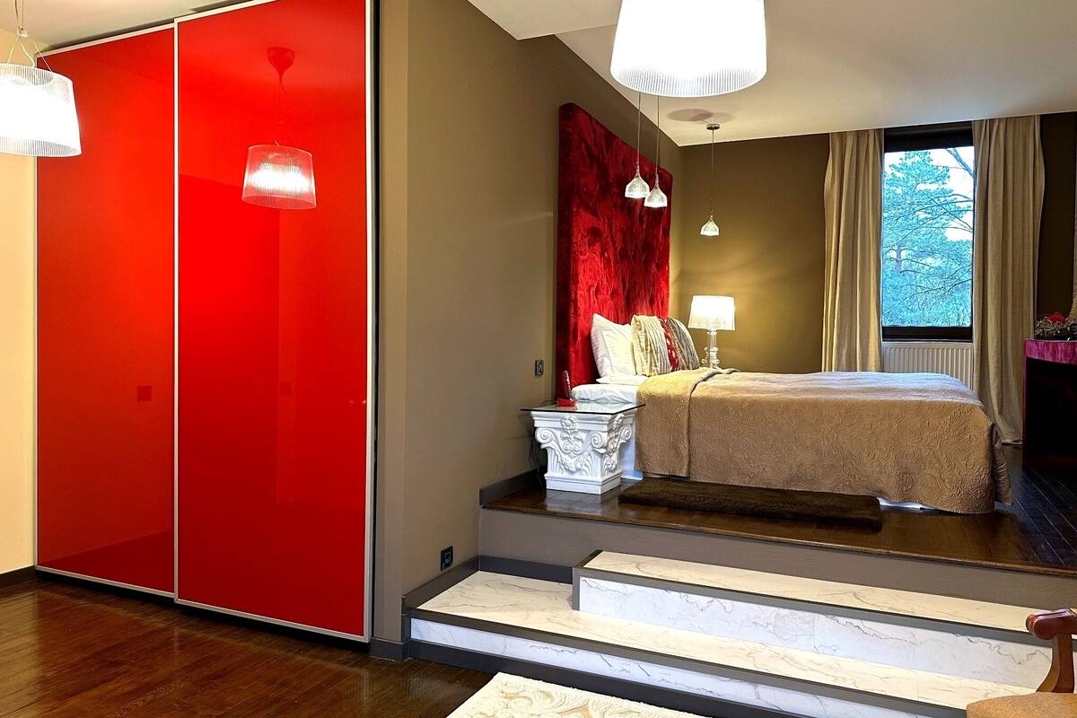 A modern bedroom with a red wardrobe, a bed with a beige bedspread, a nightstand, and a window with curtains.