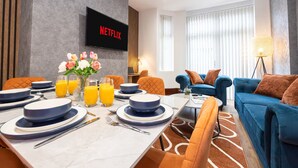 Ett modernt hotellrum med en matplats, ett vardagsrum med en blå soffa och en tv-skärm som visar ”NETFLIX”.