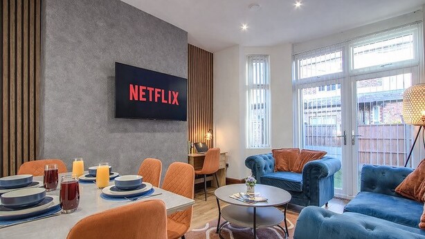 Ett modernt vardagsrum med en stor TV som visar Netflix-logotypen, en matplats med orange stolar och en blå soffgrupp.