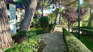 A stone pathway leading through a well-maintained garden with neatly trimmed bushes and trees, surrounded by buildings with red roofs.