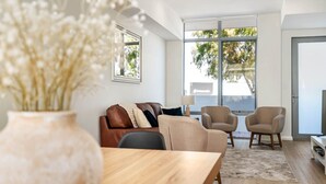 A modern living room with a wooden dining table, a brown leather sofa, two armchairs, and a large window with a view of trees. - Casa Tranquilla - Short Walk to North Coogee Beach (North Coogee)