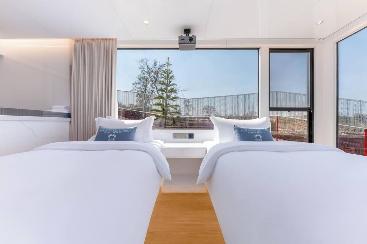 Een moderne hotelkamer met twee bedden, een groot raam en een balkon met uitzicht op een boom en gebouwen.
