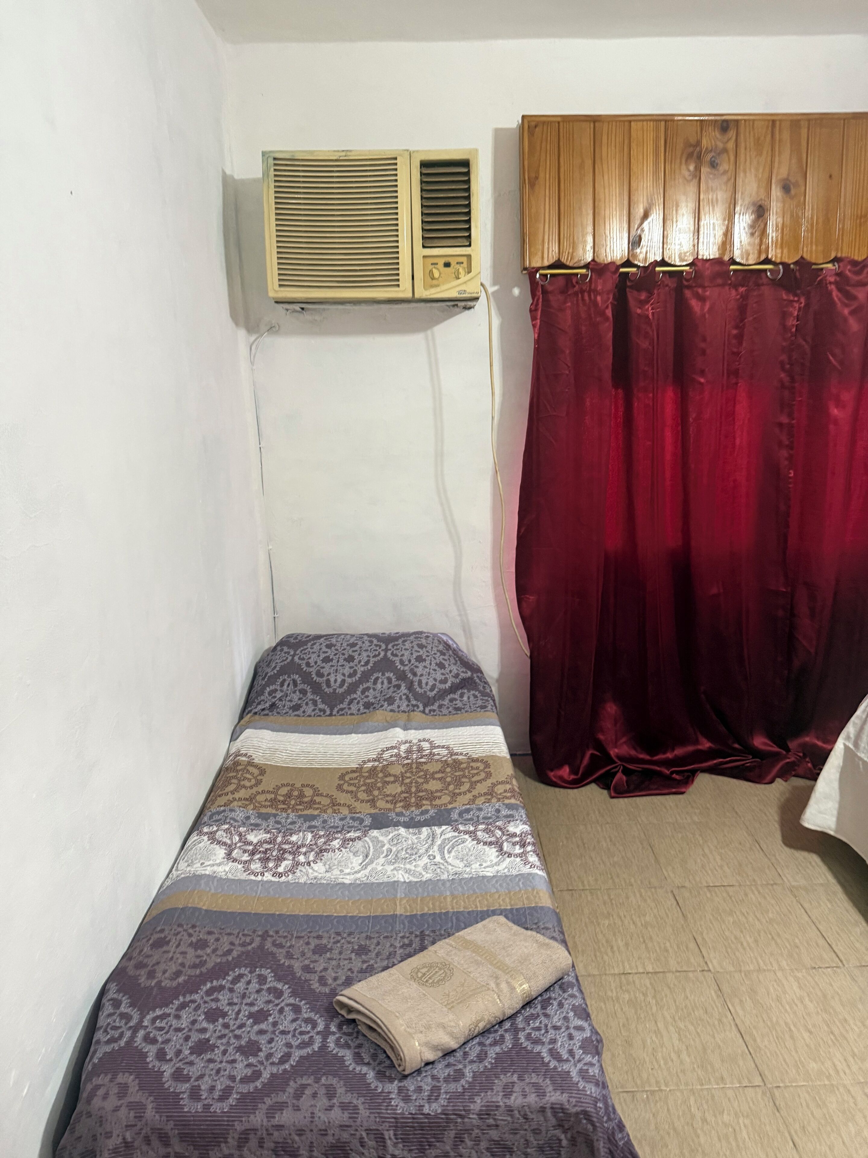 A small, single-bed room with a patterned bedspread, a red curtain, and a wall-mounted air conditioner.