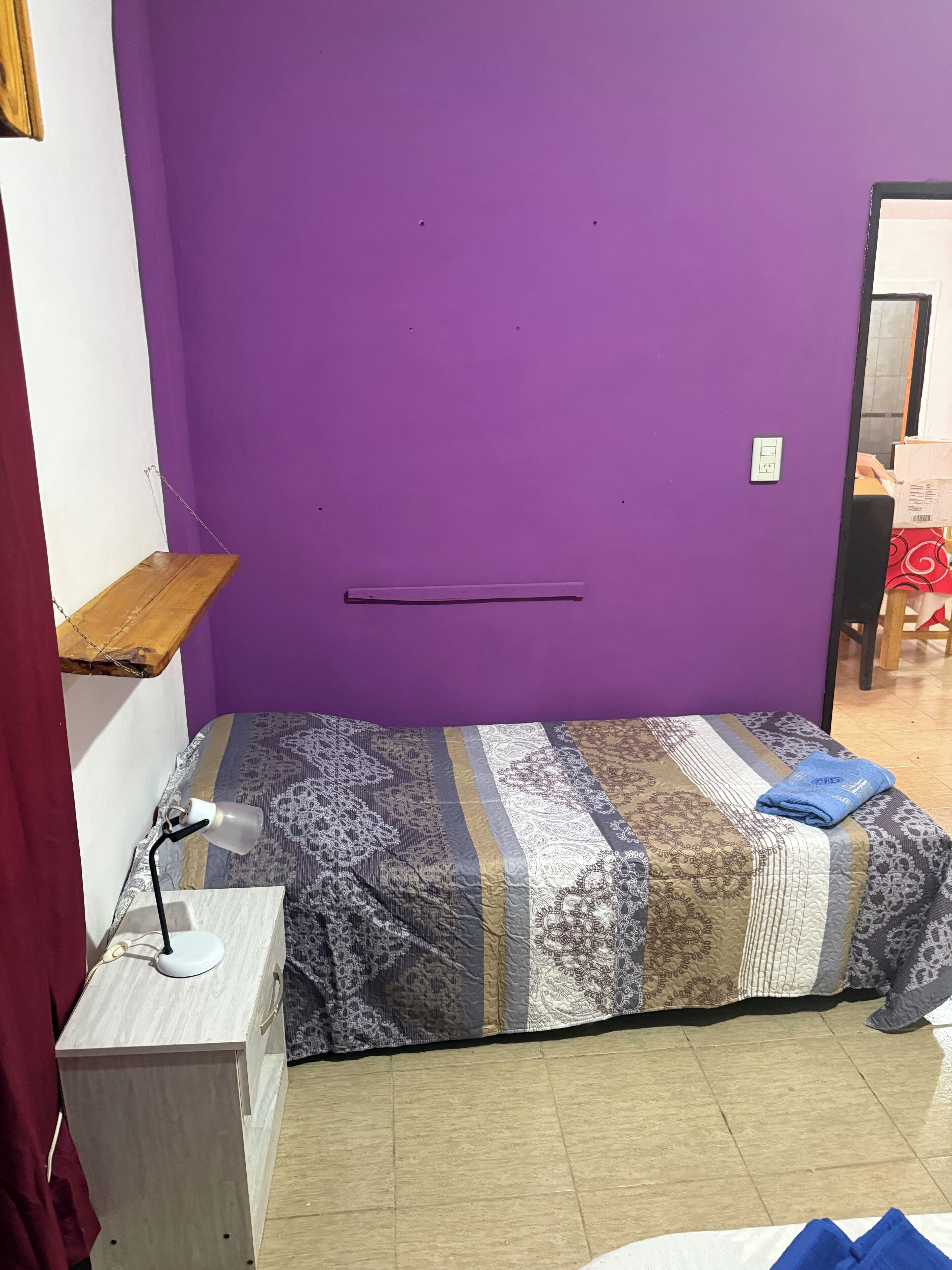 A small, purple-walled room with a bed, a desk with a lamp, and a shelf.