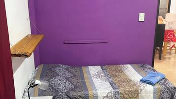 A small, purple-walled room with a bed, a desk with a lamp, and a shelf.