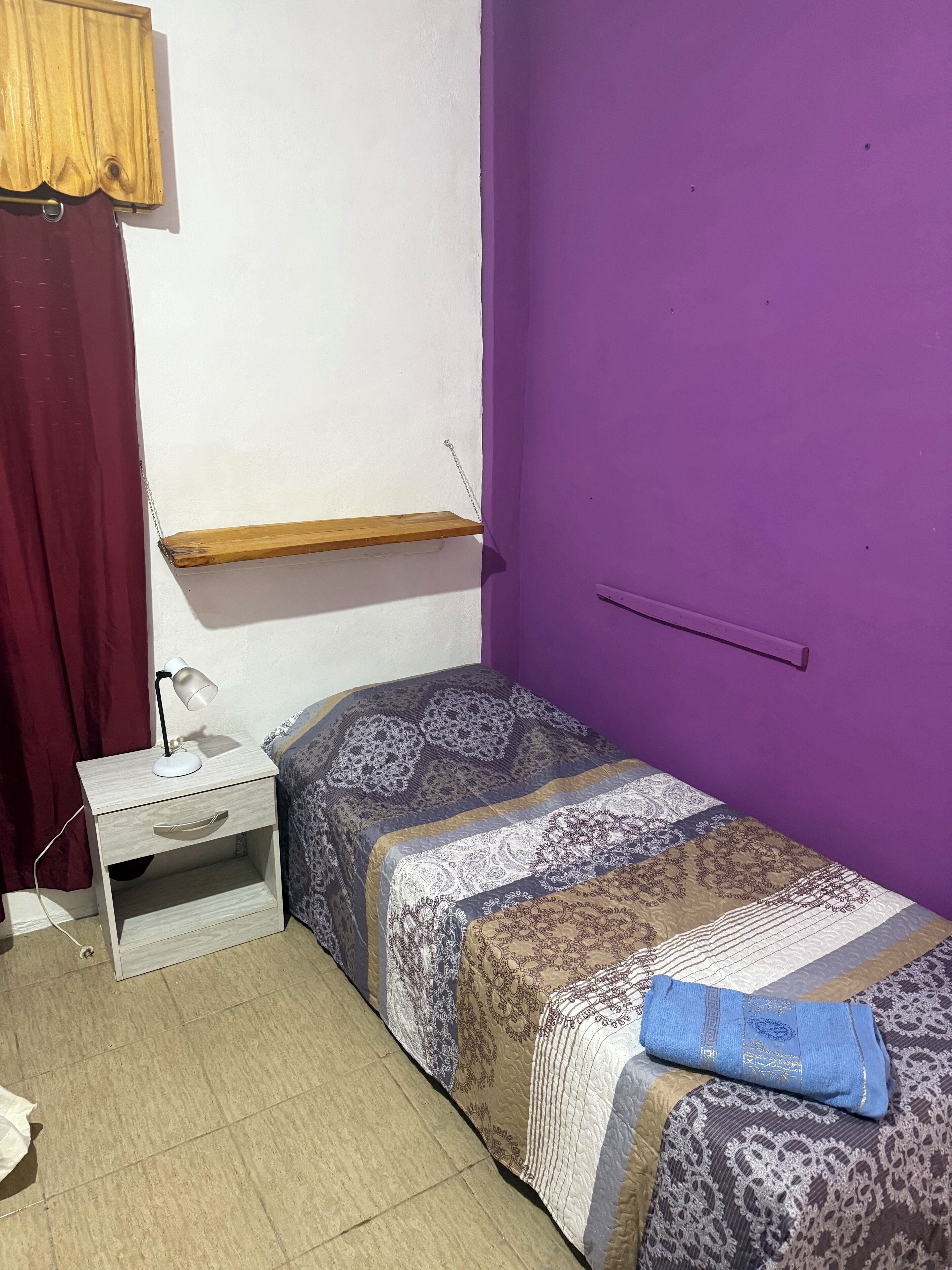 A small room with a bed, a nightstand, a purple wall, and a wooden shelf.