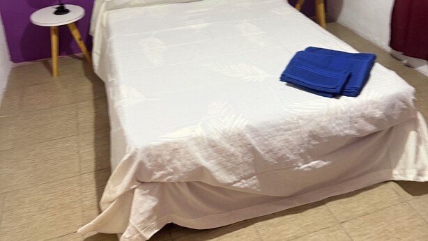 Una cama pequeña y bien arreglada con una colcha blanca y una toalla azul encima, flanqueada por dos mesitas de noche con lámparas, en una habitación con paredes moradas.