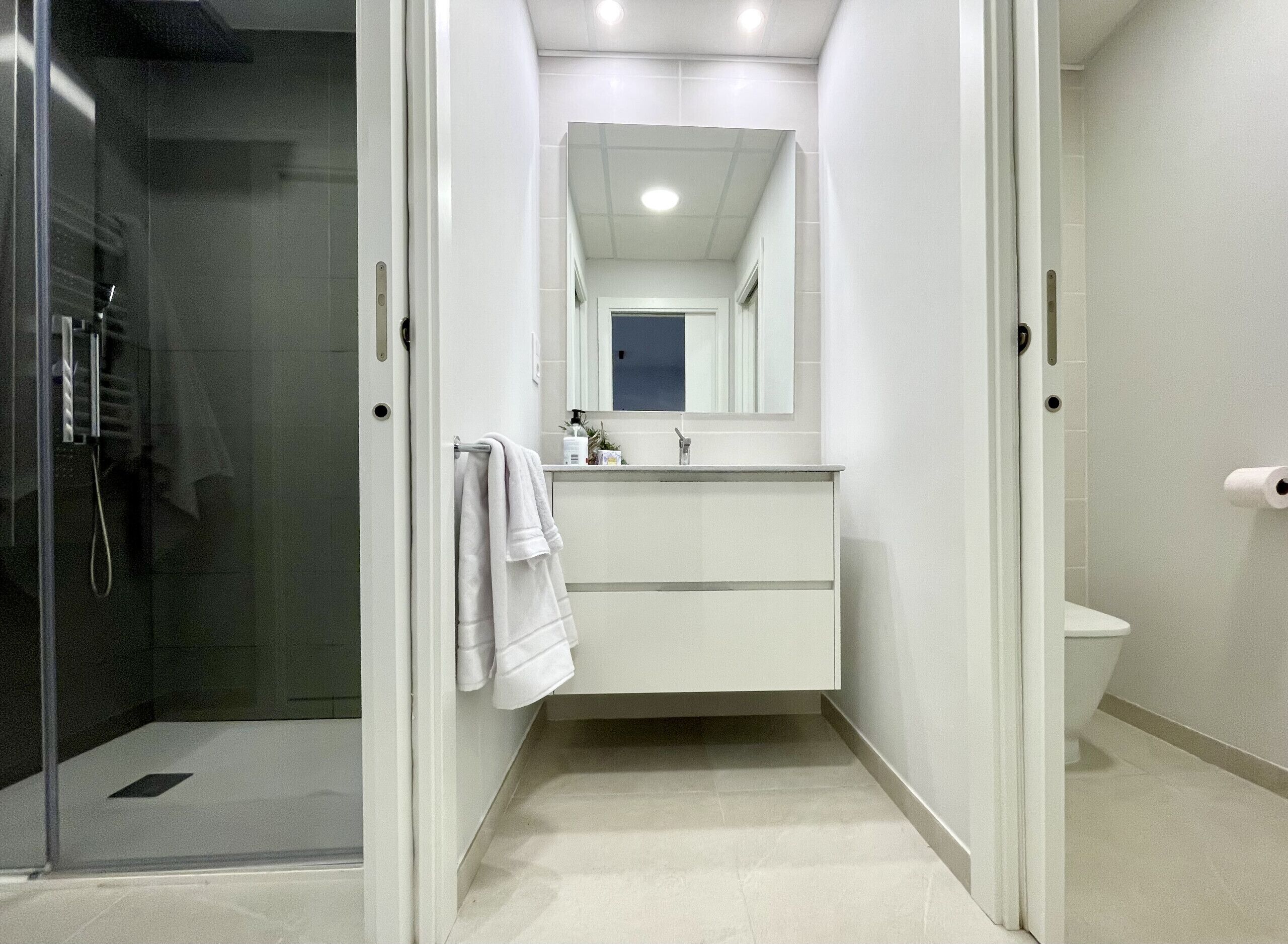 A narrow bathroom with a glass shower enclosure, a white sink with a mirror above, and a toilet.