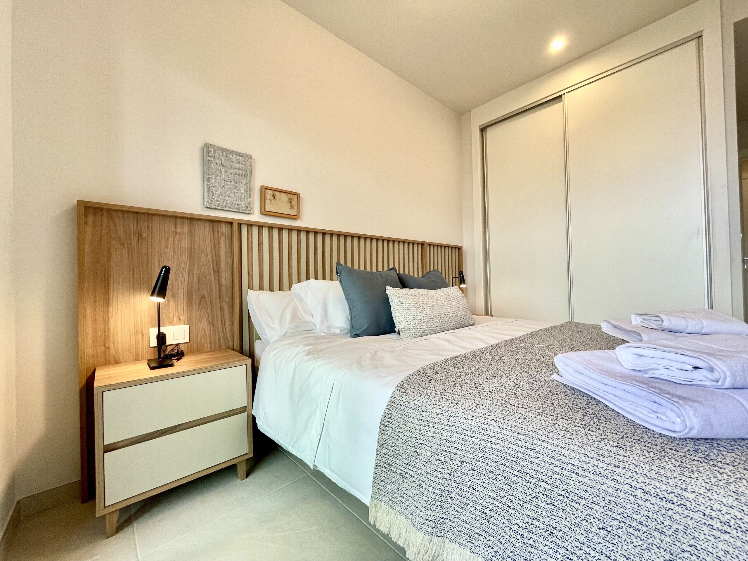 A neatly made bed with a white comforter and pillows, a wooden headboard, a bedside table with a lamp, and a folded towel on the floor.