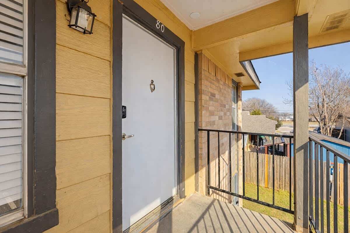 Un porche abrité avec une porte blanche portant le numéro « 80 », un petit luminaire extérieur et une vue sur un quartier résidentiel avec une clôture en bois et un bâtiment bleu.