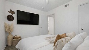 Ein Schlafzimmer mit einem Bett, einem an der Wand befestigten Fernseher, einer Tür und einem Nachttisch mit einer Vase und einer Schale.