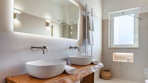 Un baño moderno con dos lavamanos grandes de color blanco, un mueble de tocador de madera, un espejo amplio, un perchero para toallas y una ventana.