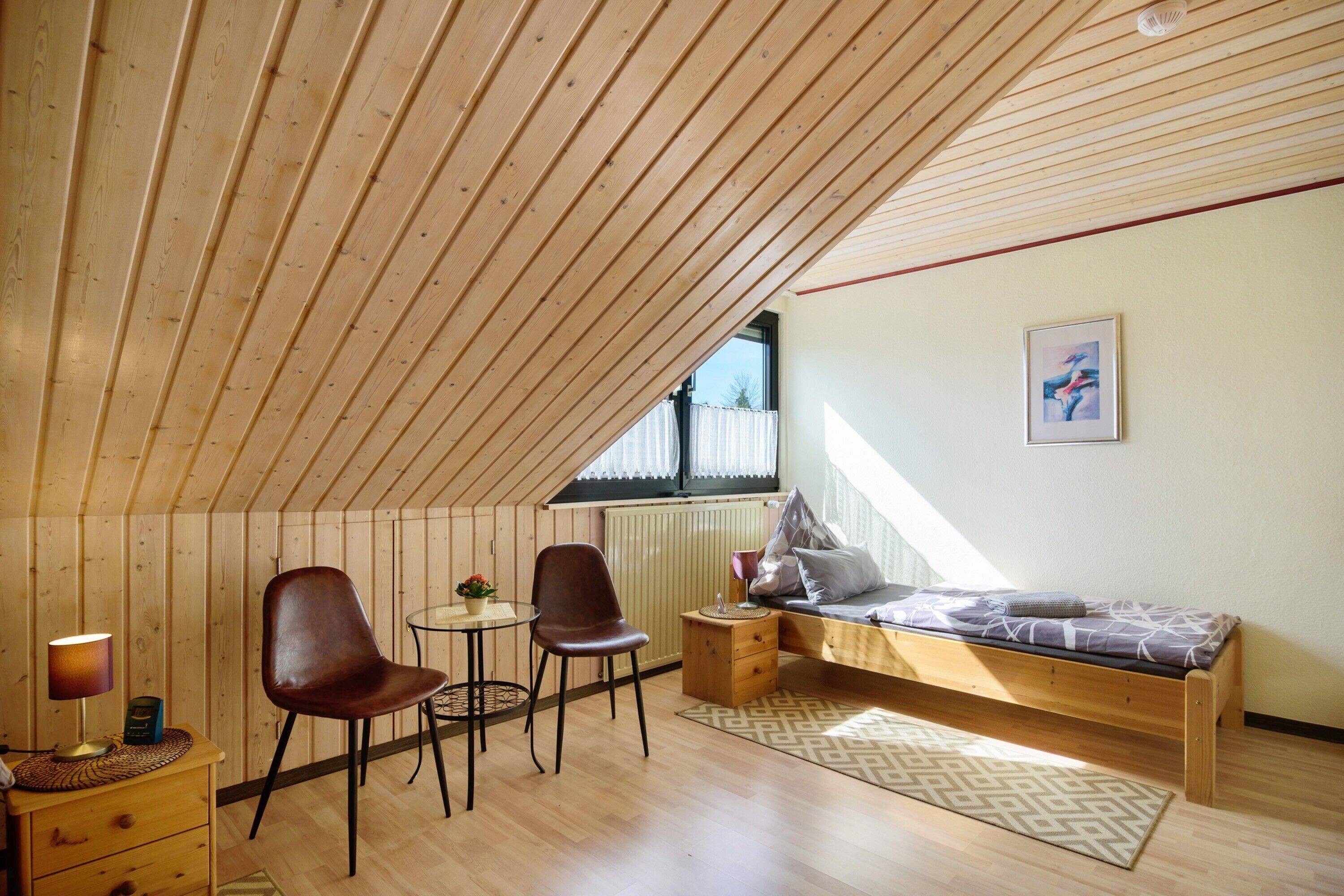 Une pièce avec un plafond en pente, un lit avec un matelas à motifs, deux chaises, une petite table avec une lampe et un radiateur.
