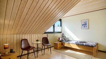Une pièce avec un plafond en pente, un lit avec un matelas à motifs, deux chaises, une petite table avec une lampe et un radiateur.