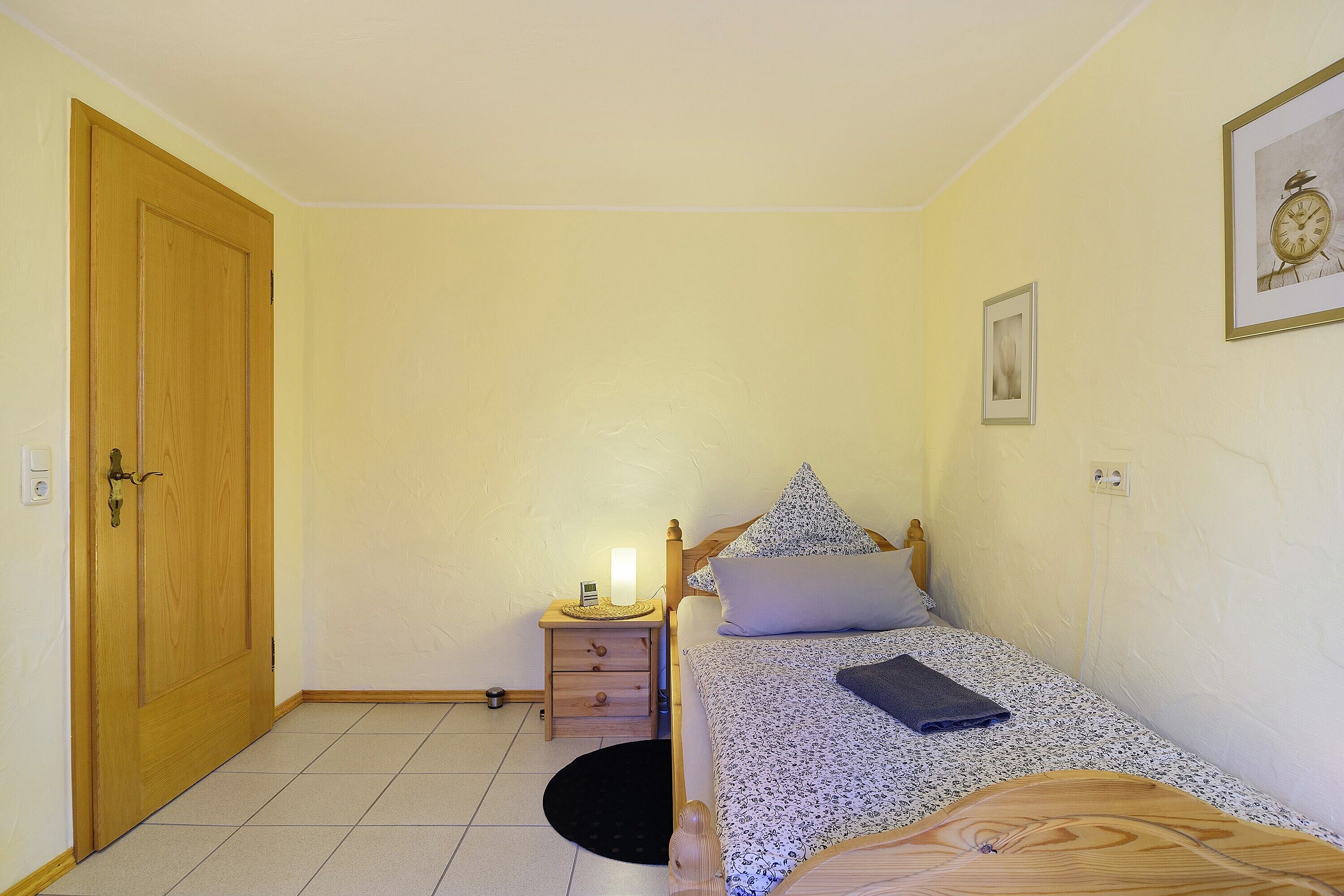 Een slaapkamer met een houten deur, een nachtkastje met een lamp, een bed met een kussen en een lijst met een afbeelding aan de muur.