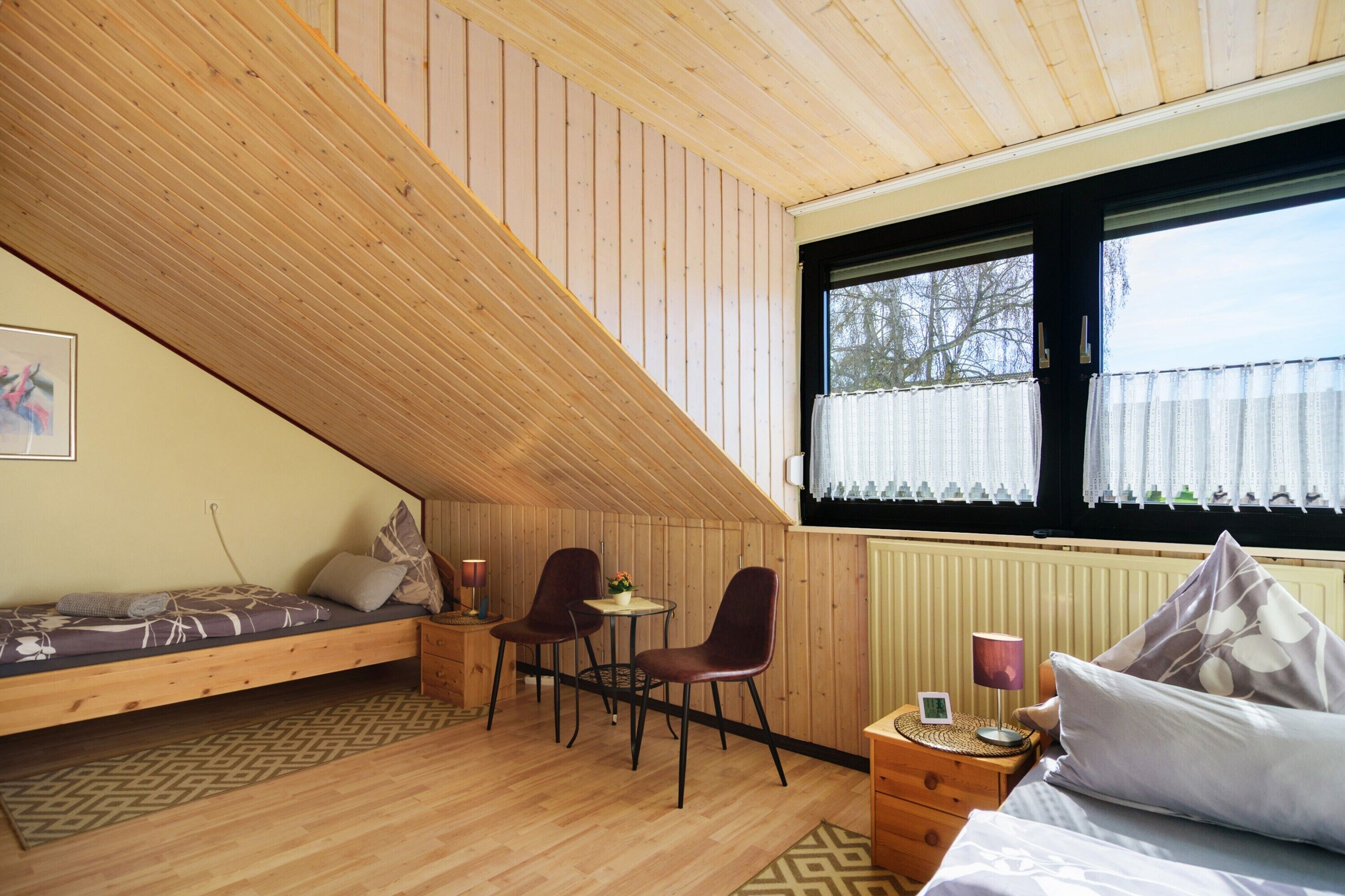 A room with a sloped ceiling, a bed, a small table with chairs, a window with curtains, and a radiator.