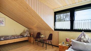 A room with a sloped ceiling, a bed, a small table with chairs, a window with curtains, and a radiator.