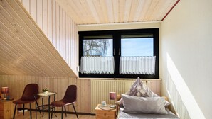 Un dormitorio con techo inclinado, una cama, dos sillas, una mesita y una ventana con cortinas.