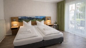 Uma cama de casal com roupa de cama branca, duas mesas de cabeceira com luminárias, um quadro na parede e vista através de uma grande janela.
