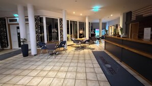 Um hall de hotel com balcão de recepção, área de estar com mesas e cadeiras, e piso de azulejos.