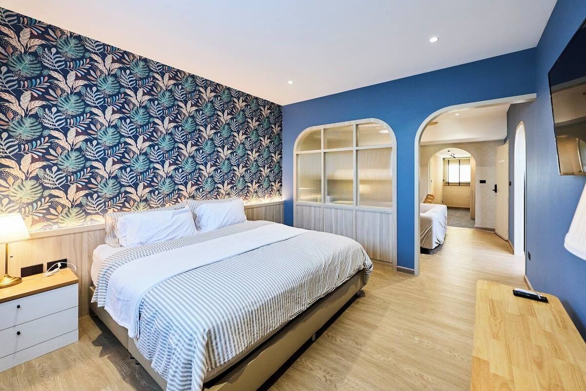 Une chambre à coucher comprenant un lit, une table de chevet, une commode, une fenêtre avec des stores et un couloir menant à une salle de bain.