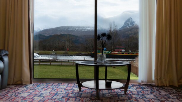 Une pièce dotée d’une grande fenêtre donnant sur une rivière, des montagnes et un autobus rouge. Une table ronde avec un vase de fleurs se trouve au premier plan.