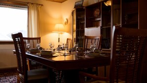 Une salle à manger avec une table en bois dressée pour quatre personnes, entourée de chaises. Une lampe se trouve sur la table, et une bibliothèque est placée sur le côté droit.
