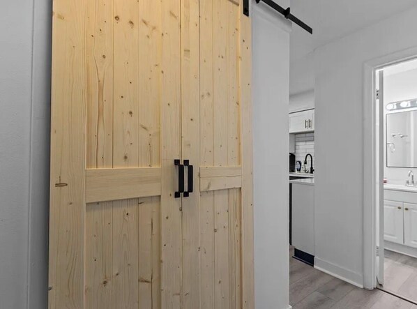 Een houten deur die leidt naar een keuken met witte kasten en een zwart aanrecht.