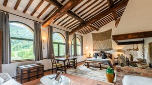 Une pièce rustique avec des poutres apparentes, une grande fenêtre avec des rideaux, une malle en bois, une table ronde, un lit avec une tête de lit et un mur de briques agrémenté d’objets décoratifs.