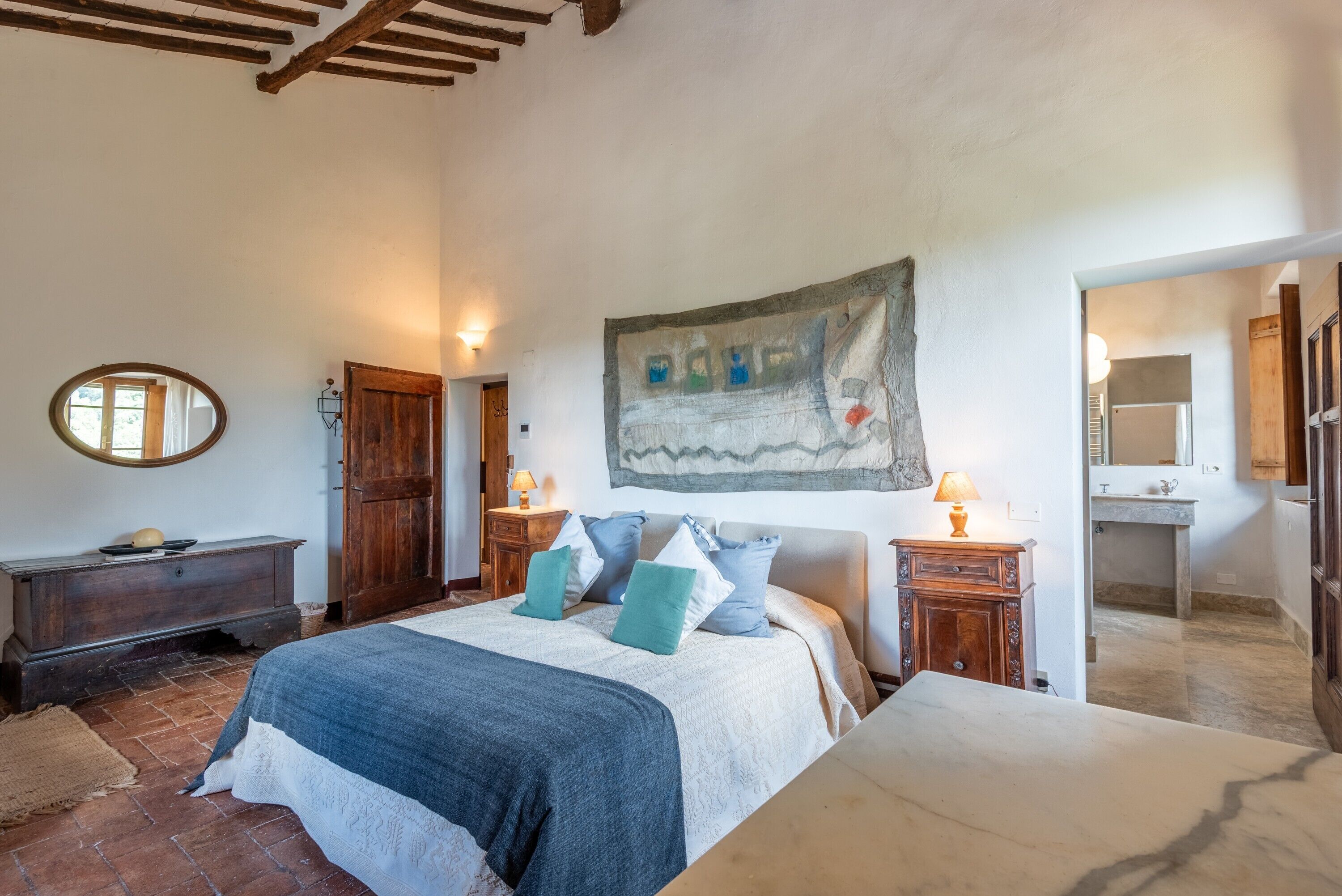 A bedroom with a bed, wooden furniture, a painting, and a bathroom visible through an open door.