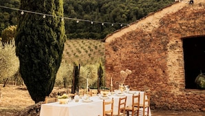 Un espace de restauration en plein air avec une table dressée pour six personnes, entouré de murs en pierre et d’un grand arbre.