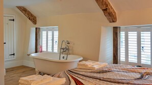 Een slaapkamer met een groot bed, een witte badkuip, houten balken in het plafond en een raam met luiken.