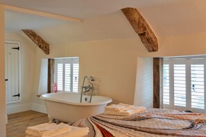 Ein Schlafzimmer mit einem großen Bett, einer weißen Badewanne, Holzbalken an der Decke und einem Fenster mit Rollläden.