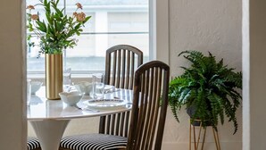 A dining area with a table set for four, surrounded by wooden chairs with striped cushions. A vase with flowers is on the table, and a potted plant is to the side.