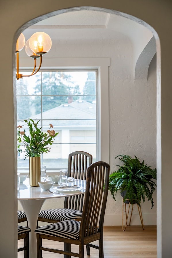 A dining area with a table set for four, surrounded by wooden chairs with striped cushions. A vase with flowers is on the table, and a potted plant is to the side.