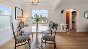 A dining area with a round table, four chairs, a chandelier, a window with a view, and a white chair in the corner.