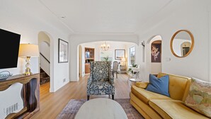 A living room with a sofa, armchair, and TV stand. There is a staircase leading to another room.