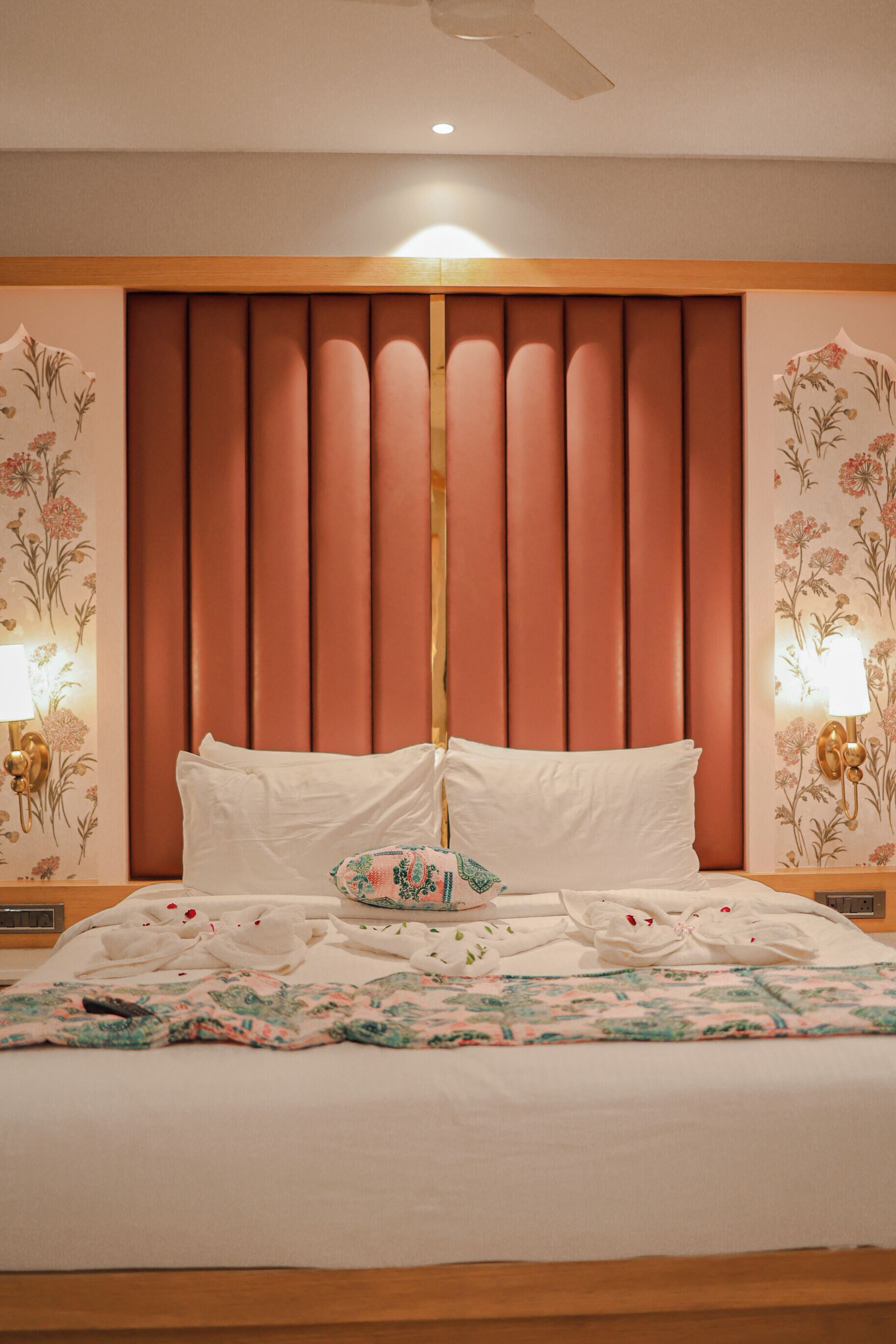 A neatly made bed with white linens and a floral-patterned blanket, a wooden headboard, and wall-mounted lamps.