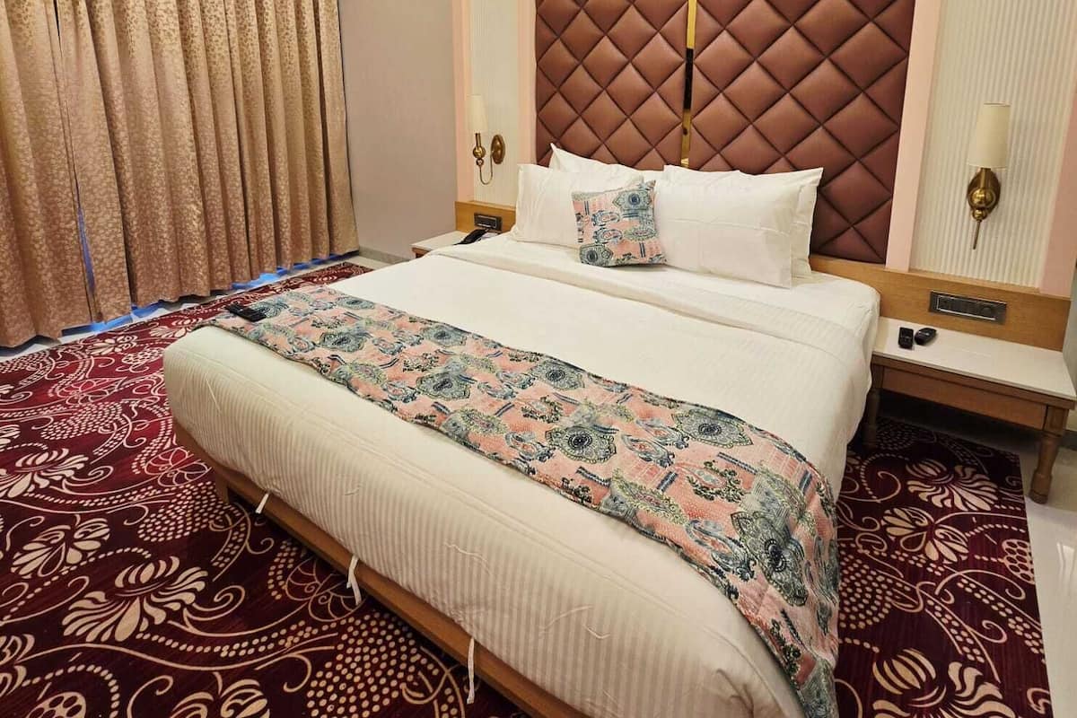 A hotel room with a large bed, a patterned carpet, a decorative headboard, and a nightstand with a lamp.