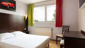 A hotel room with a bed, a desk, a chair, a window with curtains, a radiator, and a wall-mounted light fixture.