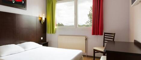 A hotel room with a bed, a desk, a chair, a window with curtains, a radiator, and a wall-mounted light fixture.