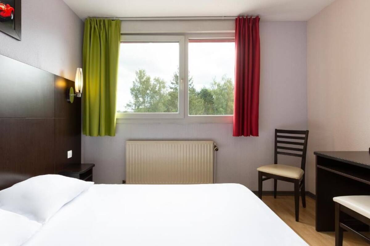 Une chambre d’hôtel avec un lit, un bureau, une chaise, un radiateur et une fenêtre avec des rideaux.